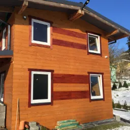 Dwukondygnacyjny dom z drewnianą elewacją w odcieniach brązu, z białymi oknami i ciemnobrązowymi akcentami, widoczny w zimowym krajobrazie z elementami budowlanymi na pierwszym planie.