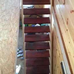 Wąskie, drewniane schody strychowe z ciemnymi stopniami w surowym wnętrzu domu w trakcie budowy, widoczne płyty OSB i drewniane panele ścienne.