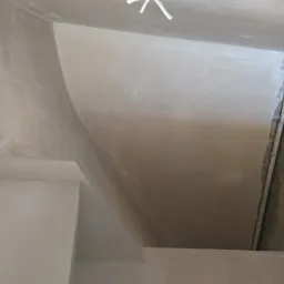 Świeżo wykończone, jasne ściany wewnętrzne z gładką powierzchnią, widoczne przejścia i narożniki, fragment sufitu z wystającymi kablami, kontrast z surową ścianą w tle.