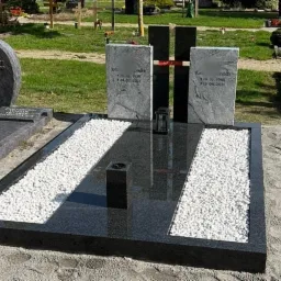 Nowoczesny, czarny pomnik z dwoma pionowymi płytami nagrobkowymi, ozdobiony białymi kamykami i metalowym krzyżem, na trawiastym cmentarzu.