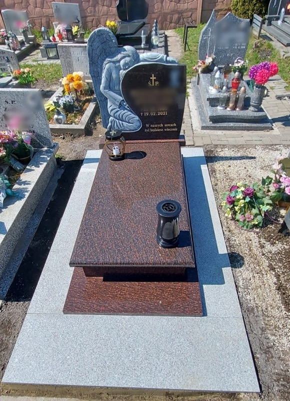 Granitowy pomnik z rzeźbą klęczącego anioła, ciemna tablica z datą i napisem, brązowo-czerwona płyta nagrobna, czarny wazon na płycie, widok na cmentarz.