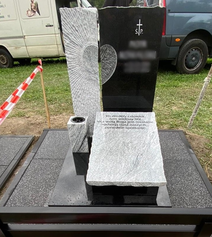 Nowoczesny, granitowy pomnik z czarnym i szarym kamieniem, z wyrytym motywem słońca i inskrypcją, ustawiony na cmentarzu, w tle biały i szary samochód dostawczy.