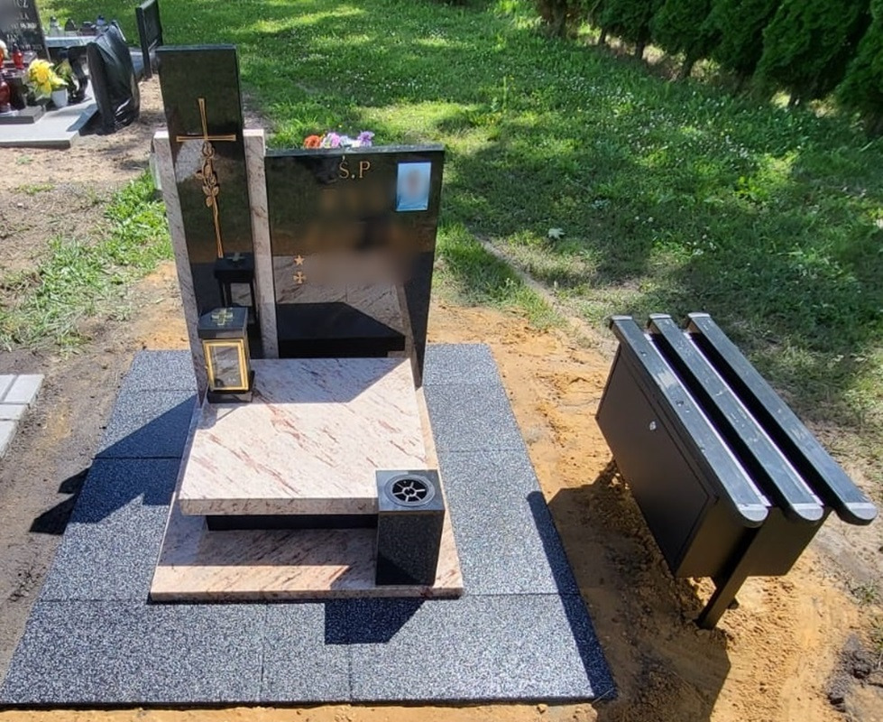 Nowoczesny, dwuelementowy pomnik z czarnego i różowego granitu, z wygrawerowanym krzyżem i różą, ustawiony na płycie z szarego granitu obok metalowej ławki cmentarnej.