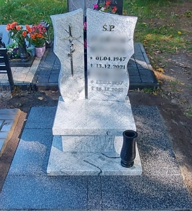 Granitowy nagrobek z krzyżem i datami, otoczony płytami chodnikowymi, z czarnym wazonem na kwiaty z boku.