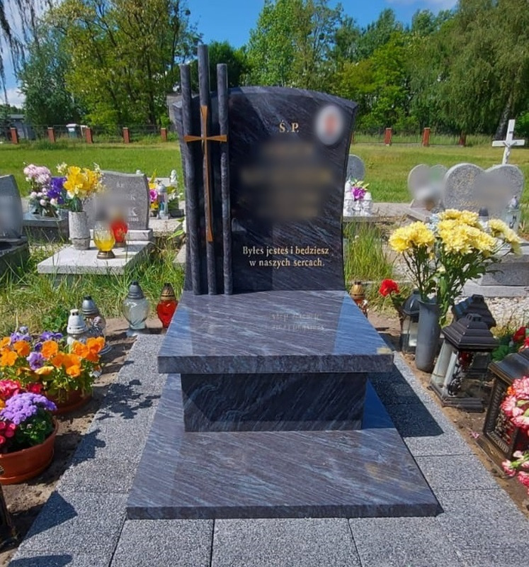 Granitowy pomnik z krzyżem na cmentarzu, otoczony kwiatami w doniczkach i zniczami, z wygrawerowanym napisem.