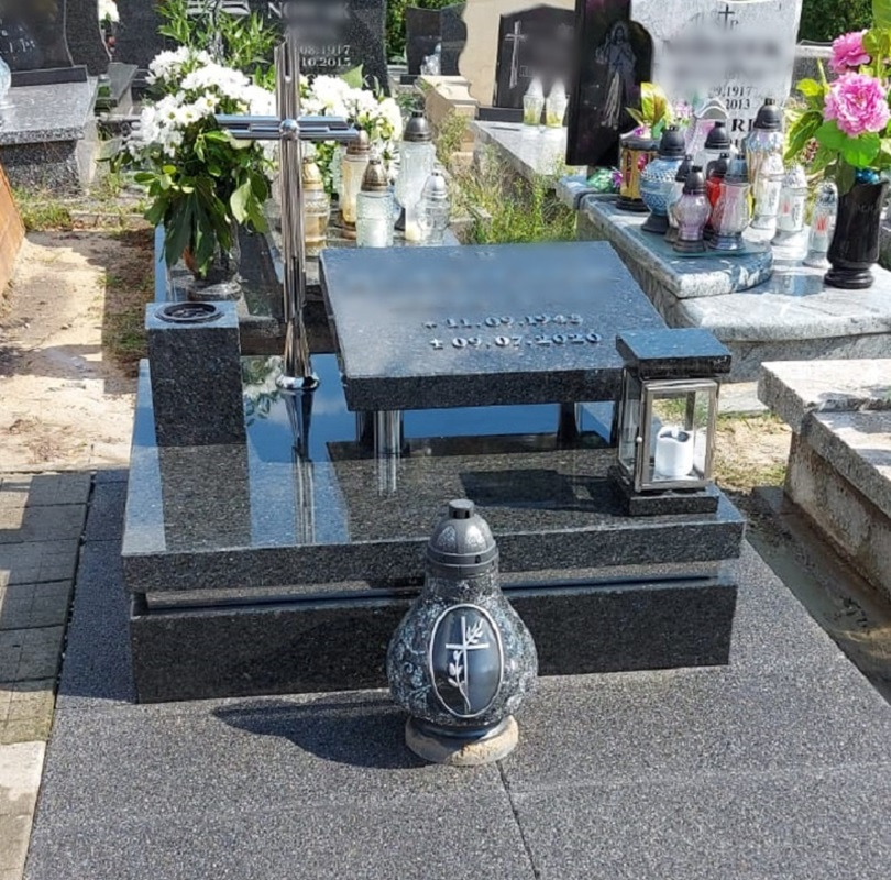 Czarny, granitowy pomnik z datami urodzin i śmierci, ozdobiony metalowym krzyżem i lampionem, otoczony innymi grobami na cmentarzu.