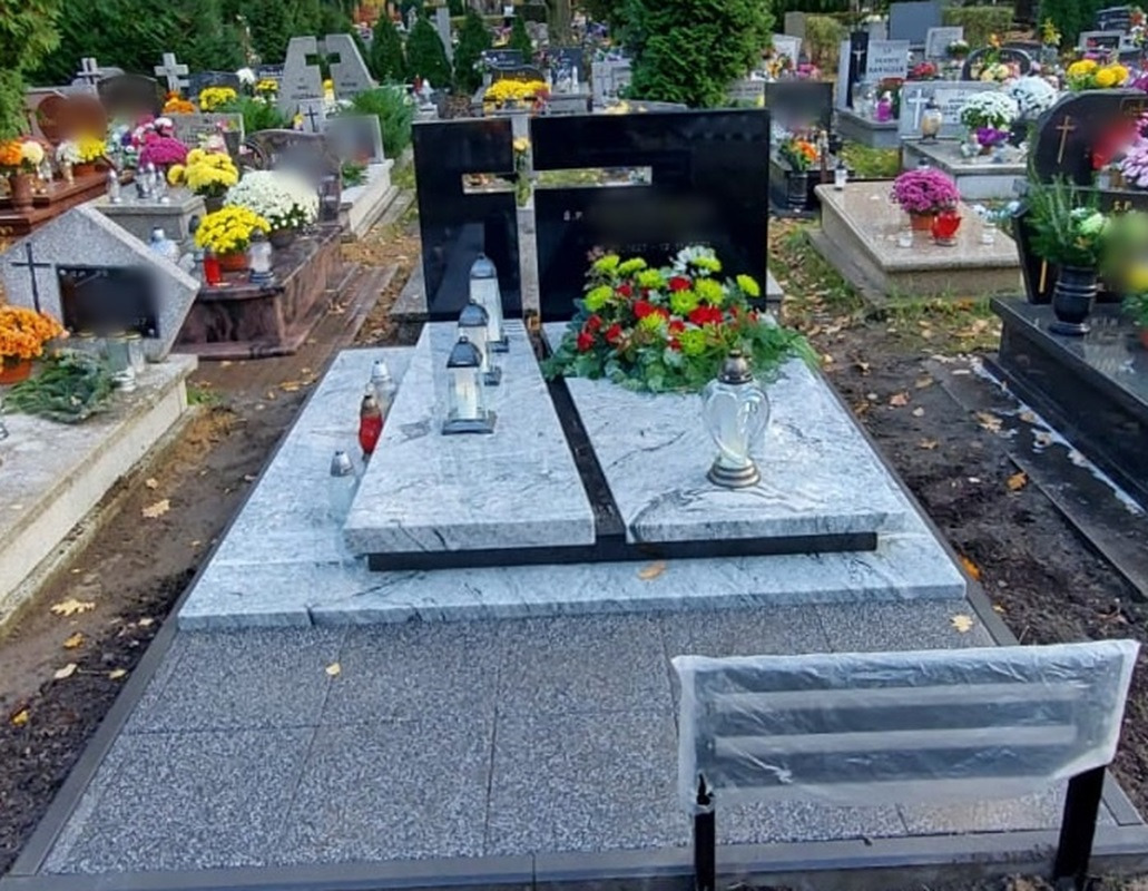 Podwójny, jasnoszary pomnik z czarnym akcentem w kształcie krzyża, udekorowany kwiatami i lampionami, na cmentarzu z widocznymi innymi grobami w tle.