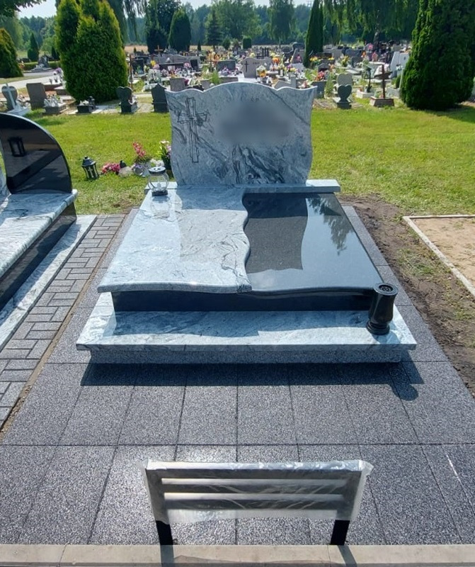 Granitowy nagrobek z ciemną płytą i jasną stelą z wyrytym krzyżem, otoczony szarą kostką brukową na cmentarzu.