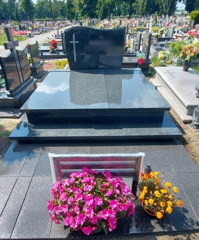 Czarny, dwupoziomowy pomnik z jasnym krzyżem na tle cmentarza, udekorowany różowymi i żółtymi kwiatami w doniczkach.