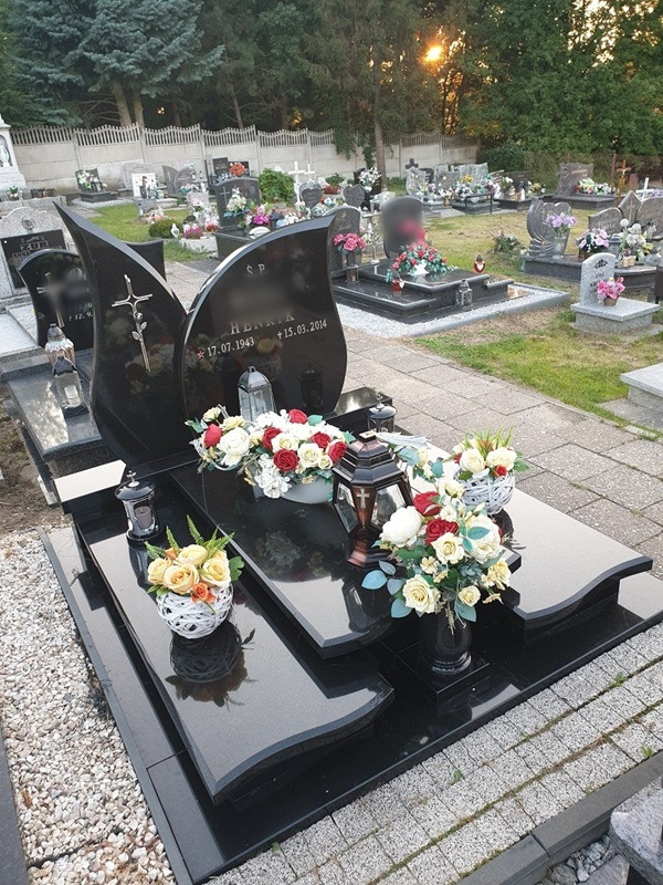 Czarny, polerowany pomnik z wygrawerowanym krzyżem i datami, udekorowany kompozycjami kwiatowymi w różnych odcieniach bieli, czerwieni i żółci, ustawiony na cmentarzu z widocznymi innymi grobami...