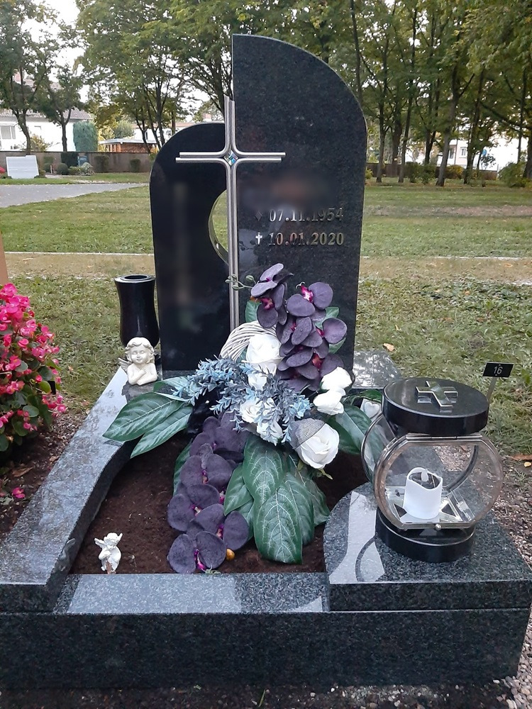 Granitowy nagrobek z krzyżem i dekoracjami kwiatowymi, zrealizowany w Niemczech. Widoczna tablica z datami oraz lampion nagrobny. Nagrobek znajduje się na cmentarzu w okolicach Rzeczyc.