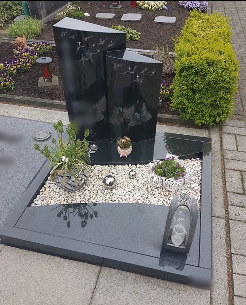 Czarny, nowoczesny pomnik z dwoma pionowymi elementami o asymetrycznych kształtach, zdobiony motywem gwiazd, otoczony białym żwirem, roślinami w doniczkach i metalowymi kulami, z lampionem...