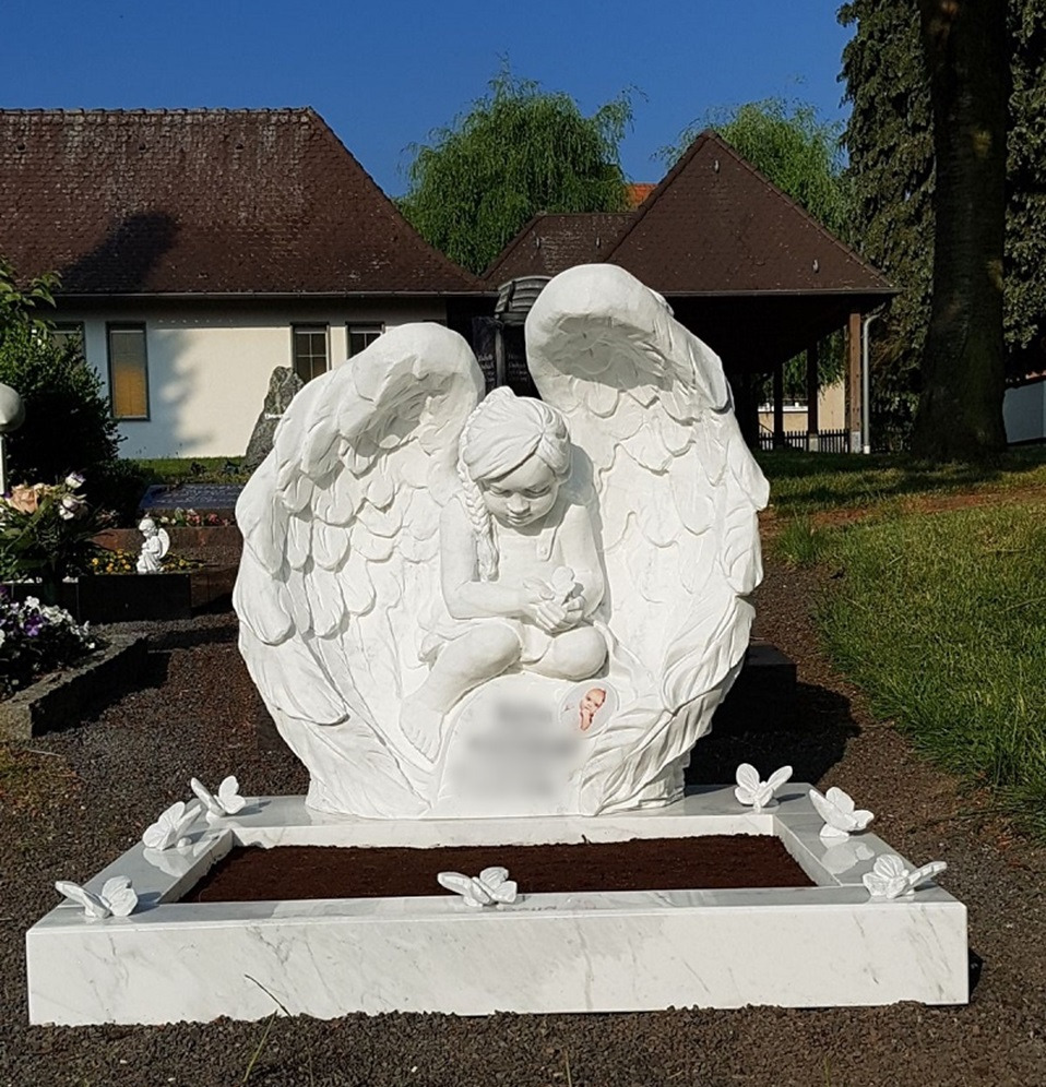 Biały pomnik z rzeźbą anioła z rozłożonymi skrzydłami, siedzącego na podstawie z motylami, na tle cmentarza z zielenią i budynkami.