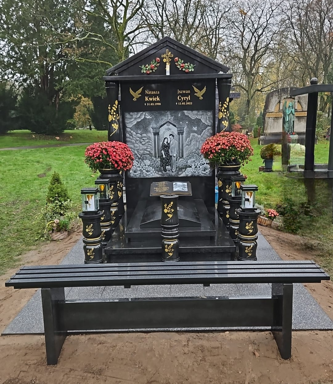 Czarny, ozdobny pomnik z fotografią pary, kwiatami w donicach i ławką na cmentarzu, z inskrypcjami Natasza KwieK i Iszwan Cyryl.