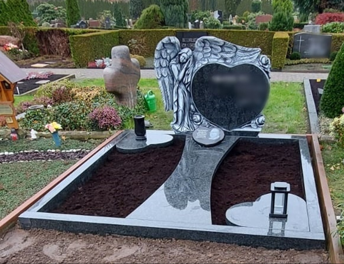 Granitowy nagrobek z rzeźbą anioła i sercem w Niemczech, otoczony ziemią przygotowaną pod nasadzenia.