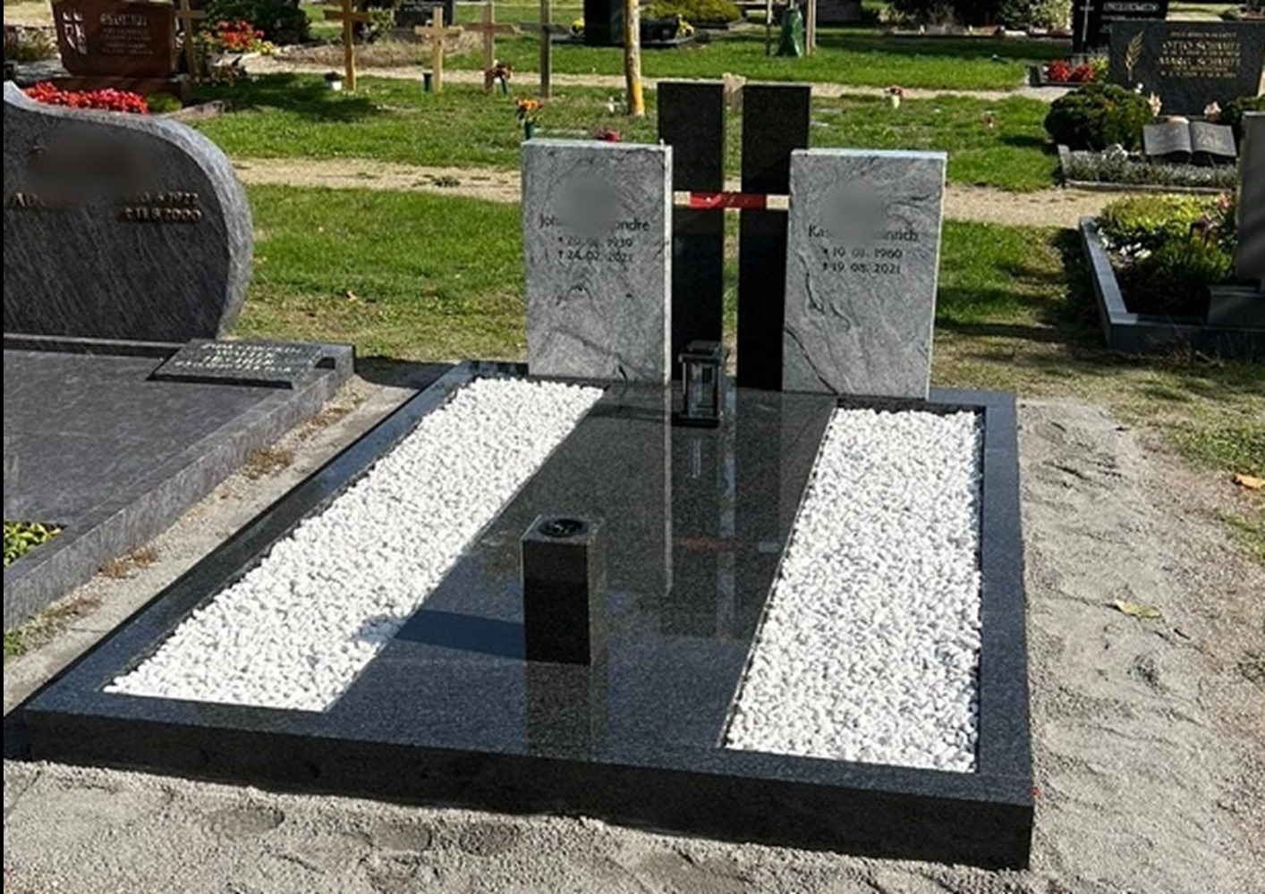 Nowoczesny, czarny pomnik z dwoma pionowymi płytami nagrobkowymi, ozdobiony białymi kamykami i metalowym krzyżem, na trawiastym cmentarzu.