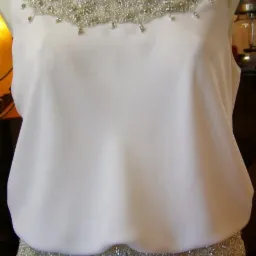 Biała, bezrękawnikowa suknia z ozdobnym, srebrnym zdobieniem z koralików wokół dekoltu i w pasie, prezentowana na manekinie.