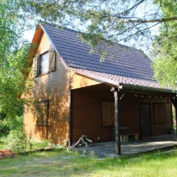 Brunatny dom szkieletowy z zadaszonym tarasem, otoczony zielenią. Widoczne drewniane okiennice i dekoracyjne oświetlenie tarasu. Ujęcie w słoneczny dzień.