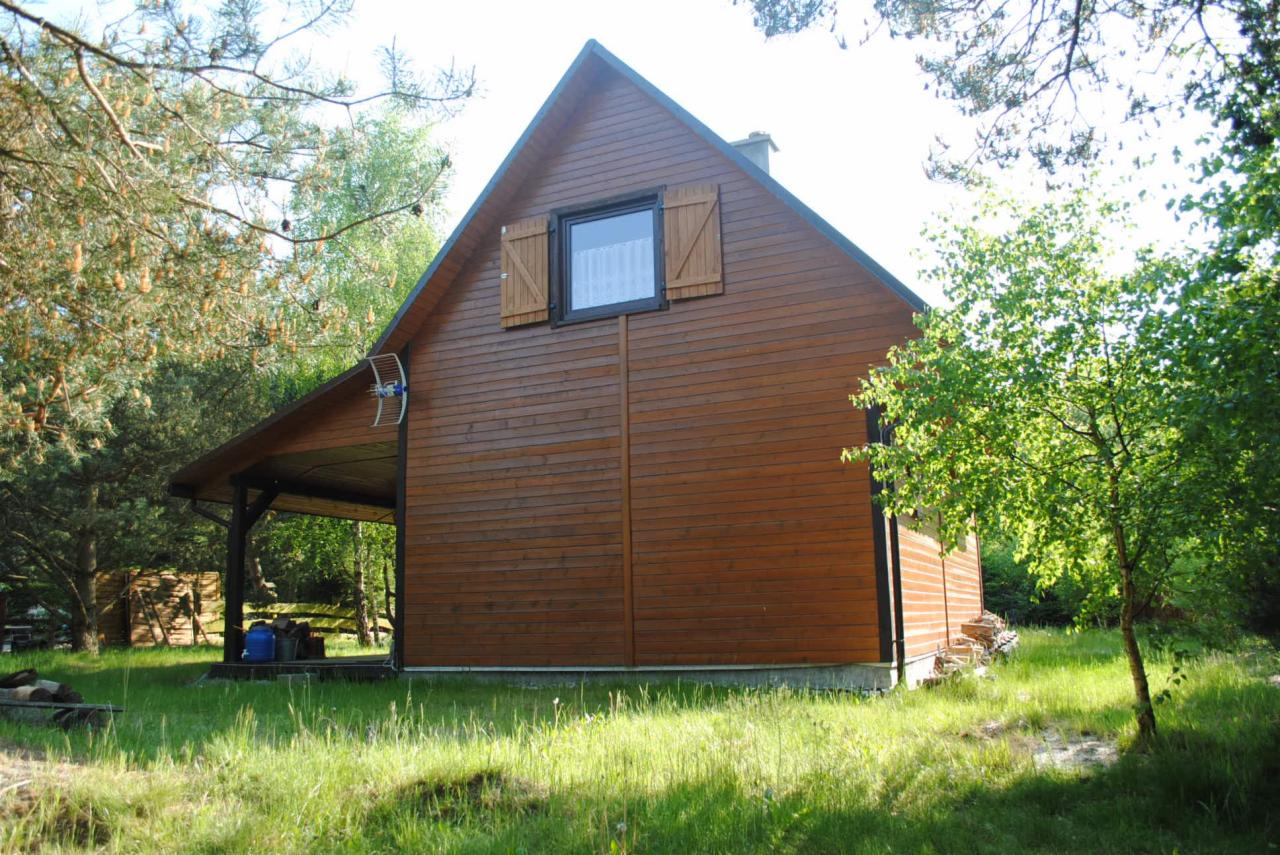 Brązowy dom szkieletowy z drewnianymi okiennicami, widoczny z zewnątrz, otoczony zielenią drzew i trawy. Antena satelitarna na ścianie.