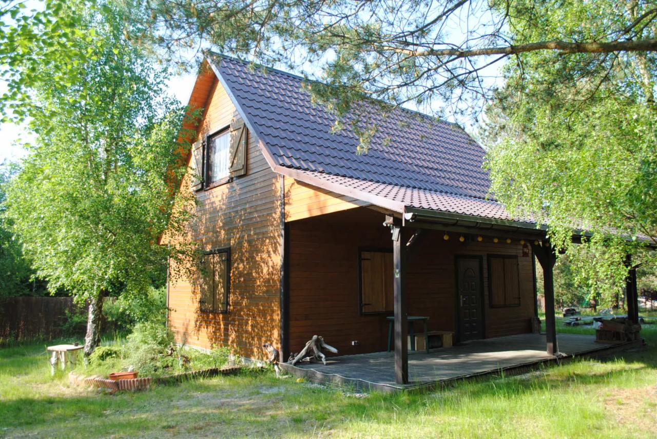 Brunatny dom szkieletowy z zadaszonym tarasem, otoczony zielenią. Widoczne drewniane okiennice i dekoracyjne oświetlenie tarasu. Ujęcie w słoneczny dzień.