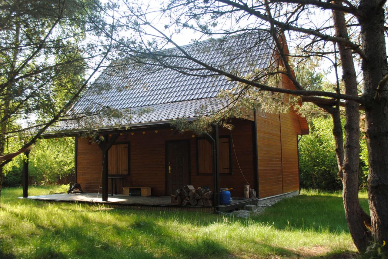 Drewniany domek letniskowy z zadaszonym tarasem, otoczony zielenią drzew. Na tarasie widoczne drewno na opał i niebieska butla gazowa. Dach pokryty szarą dachówką.