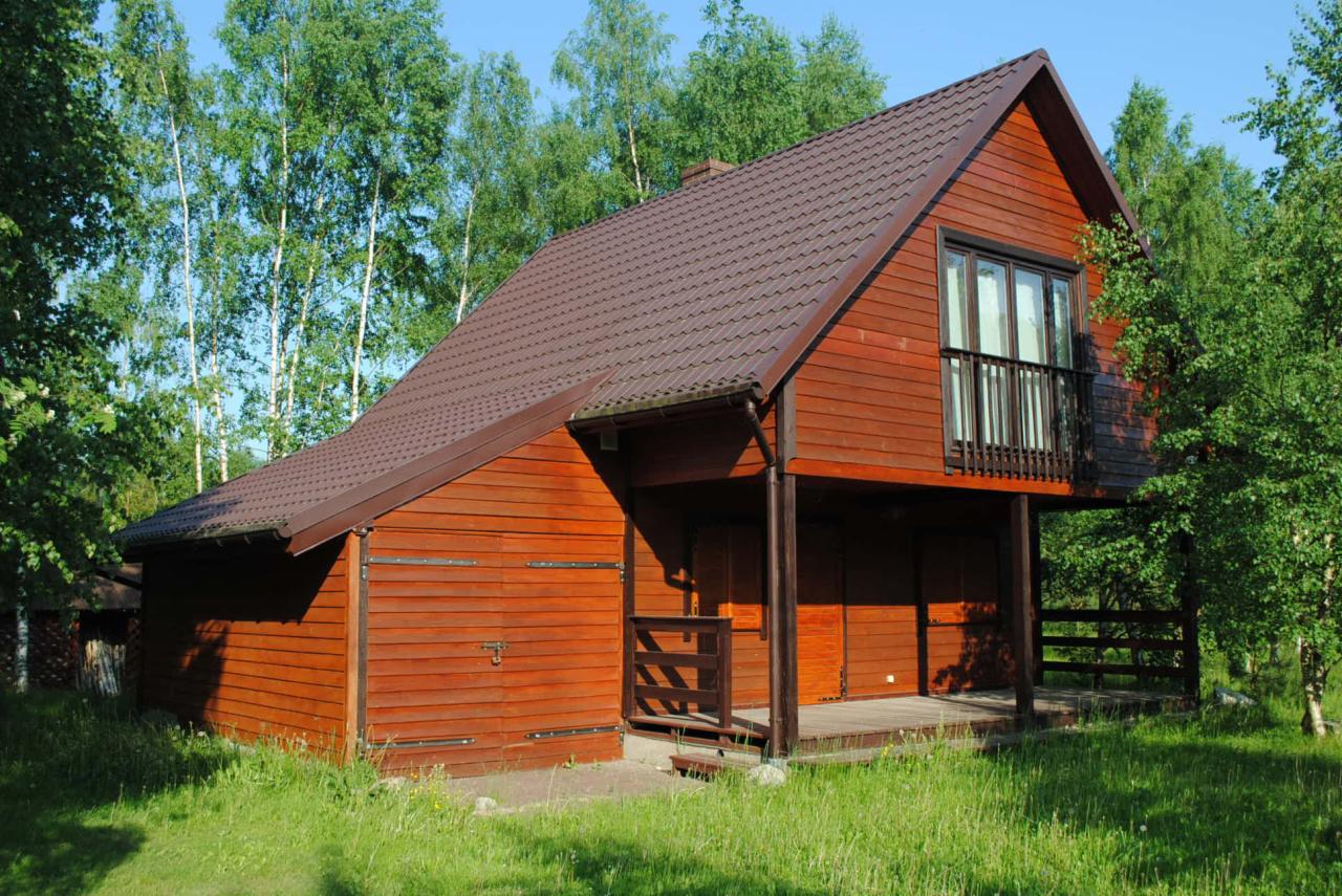 Drewniany dom z brązowym dachem i małym balkonem na tle drzew. Budynek z drewnianą elewacją, widoczny na tle zieleni i błękitnego nieba.