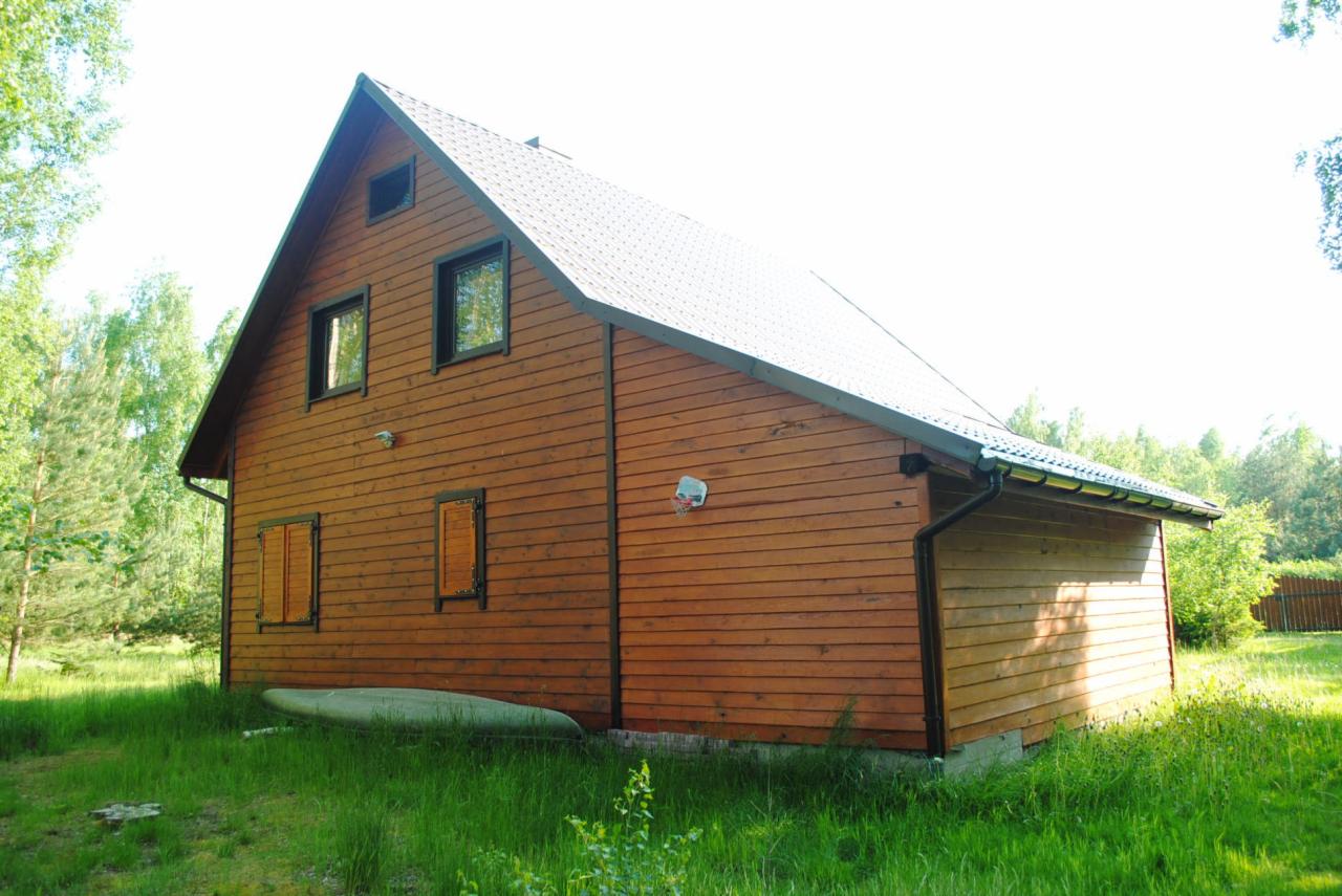 Drewniany dom szkieletowy z szarym dachem i zielonym otoczeniem. Na trawie leży kajak, a na ścianie wisi kosz do koszykówki. Widok z dołu, słoneczny dzień.