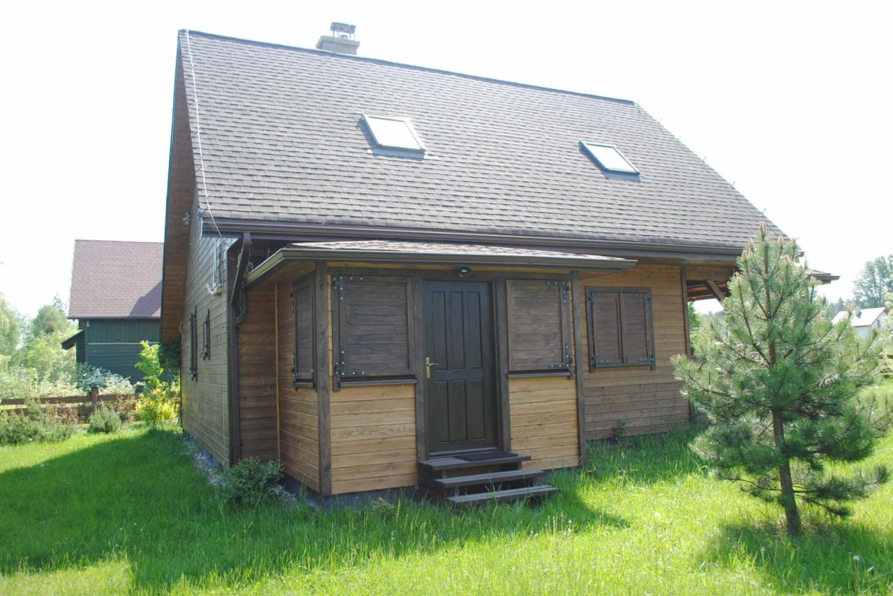 Dom szkieletowy z drewnianą werandą i brązowymi okiennicami na tle zielonego trawnika. Dach pokryty gontem bitumicznym z dwoma oknami połaciowymi.