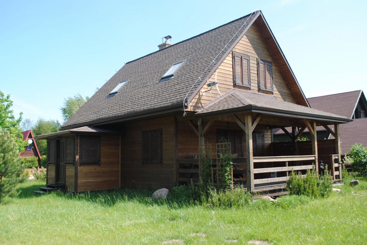 Drewniany dom z brązowym dachem i drewnianymi okiennicami, zadaszony taras z drewnianą balustradą, widok z zewnątrz w słoneczny dzień.