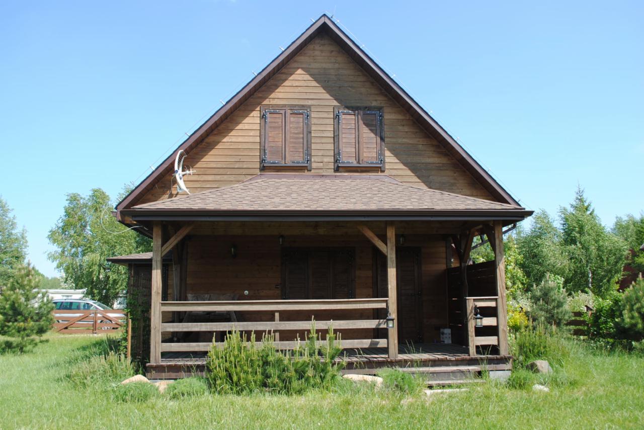 Drewniany dom szkieletowy z zadaszonym tarasem i brązowym dachem. Widoczna antena satelitarna i okiennice. Dom otoczony zielenią w słoneczny dzień.