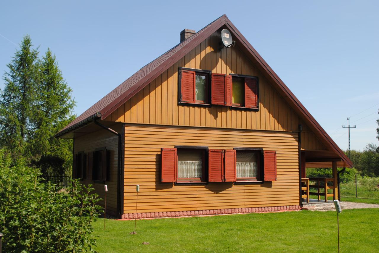 Drewniany dom z czerwonymi okiennicami w Choczewo. Elewacja z desek, brązowy dach i zadbany trawnik. Widoczna antena satelitarna i komin.