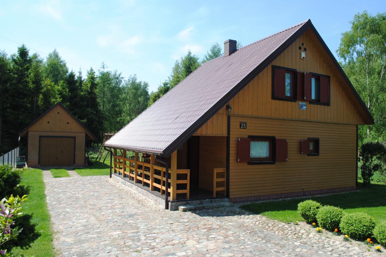 Żółty dom szkieletowy z brązowym dachem i okiennicami, widoczny z kamiennej ścieżki. Na posesji znajduje się również garaż.