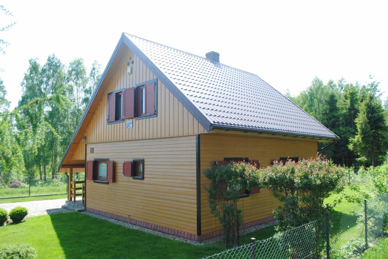 Żółty dom szkieletowy z brązowym dachem i okiennicami, otoczony zielenią. Widoczny fragment ganku i numer domu '25'.