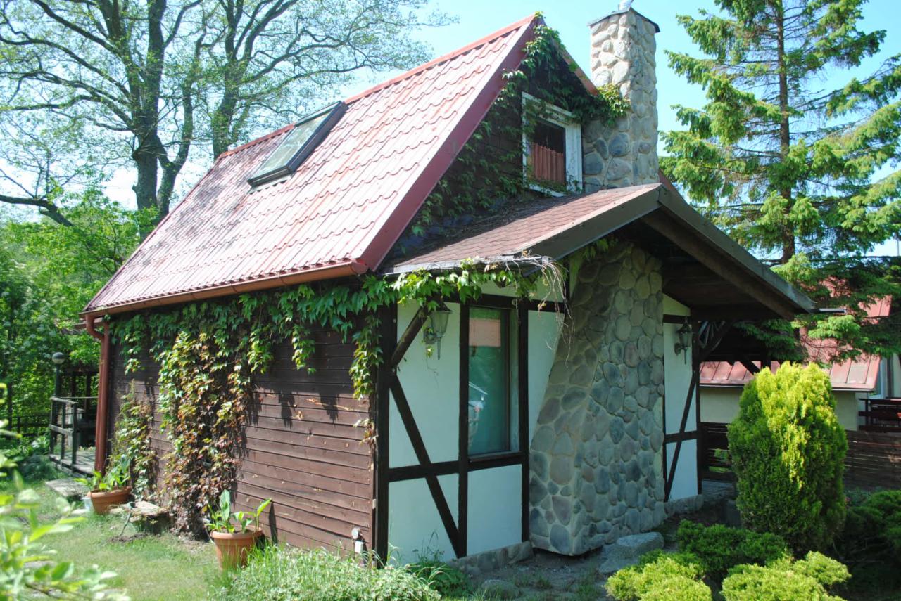 Urokliwy dom z czerwonym dachem, kamiennym kominem i drewnianą elewacją, częściowo porośnięty bluszczem. Widoczne okno dachowe i elementy konstrukcji szkieletowej.