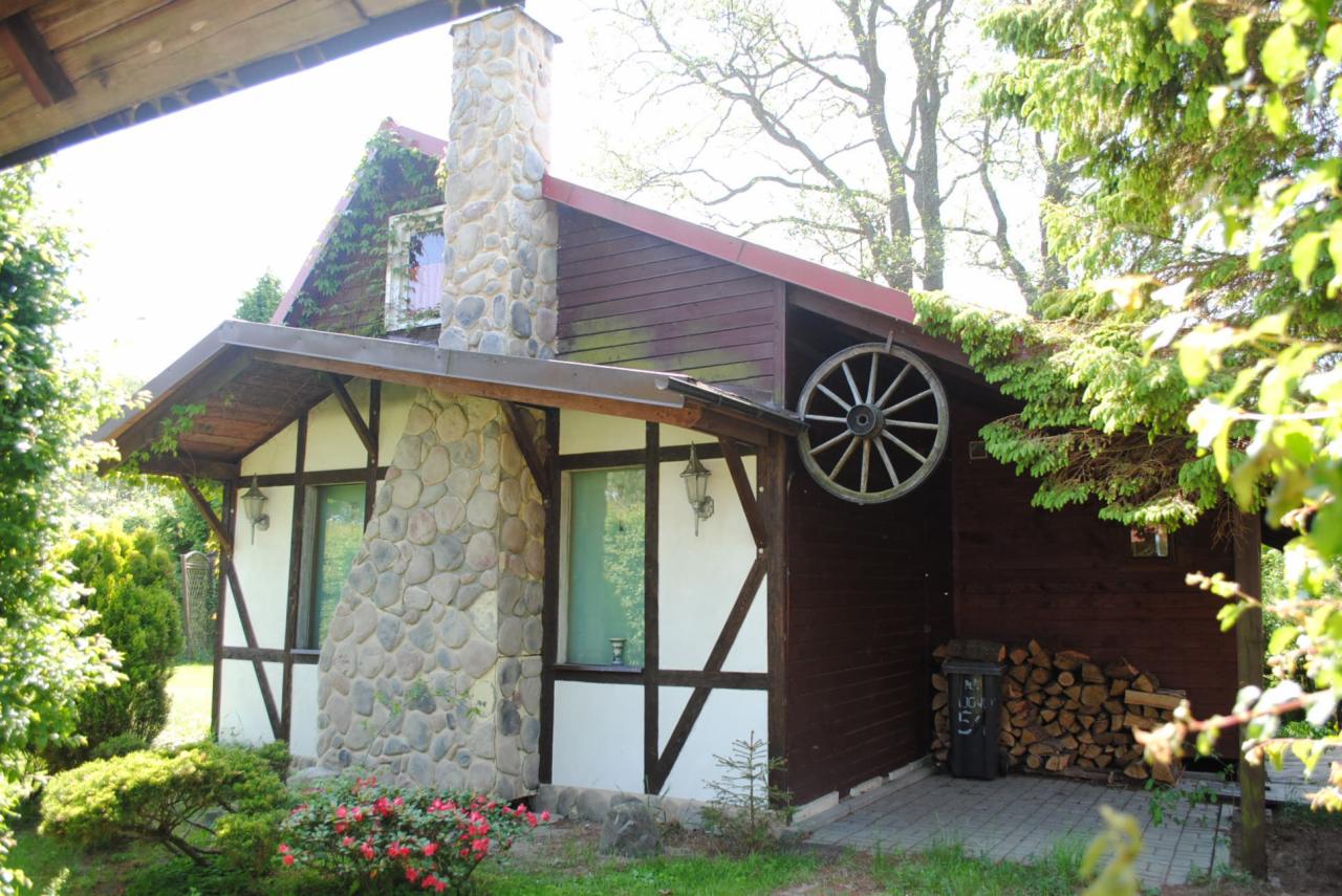 Dom z brązową elewacją, kamiennym kominem i dekoracyjnym kołem wozu na ścianie. Widoczne drewno na opał i bujna roślinność wokół.