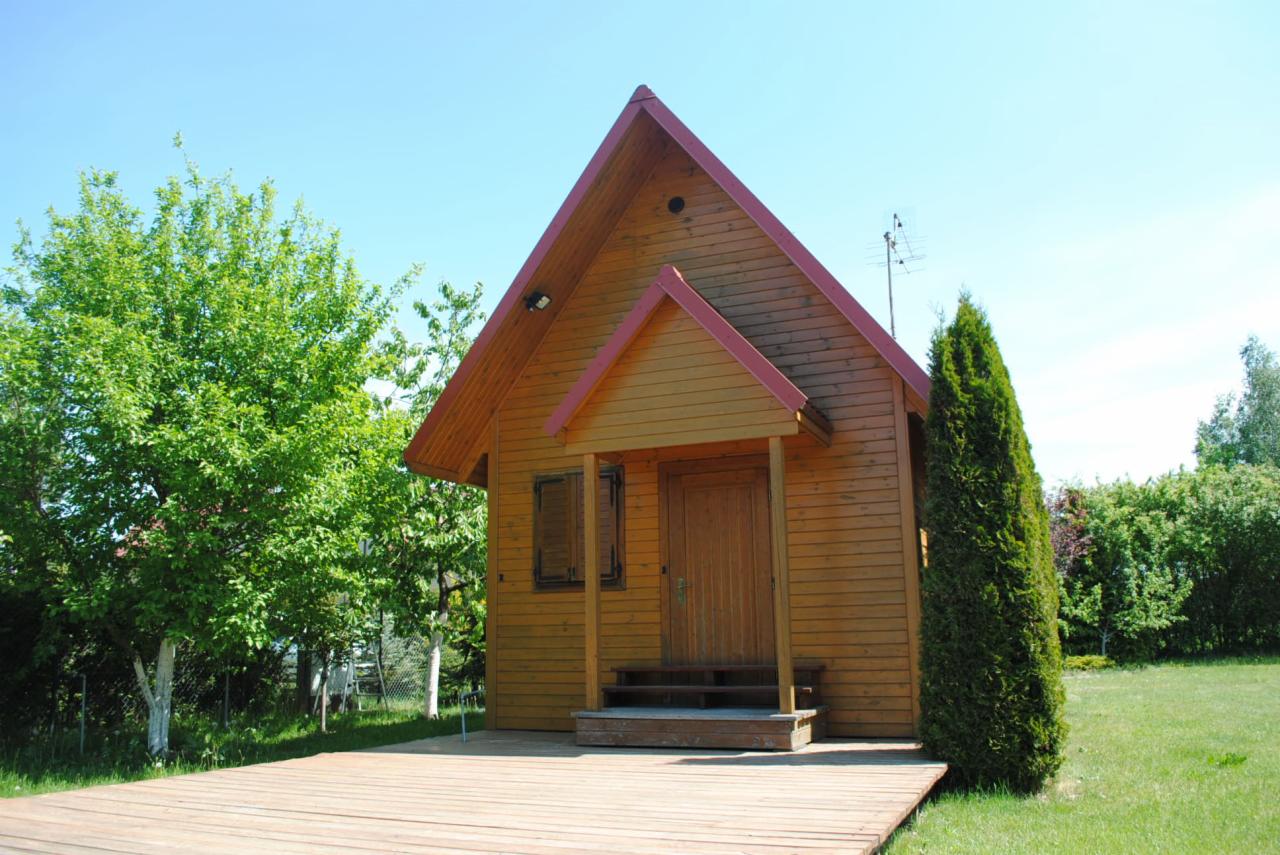 Drewniany domek z czerwonym dachem i tarasem, otoczony zielenią. Widoczne drzwi wejściowe, okno z okiennicą i antena na dachu. Jasne, słoneczne ujęcie.