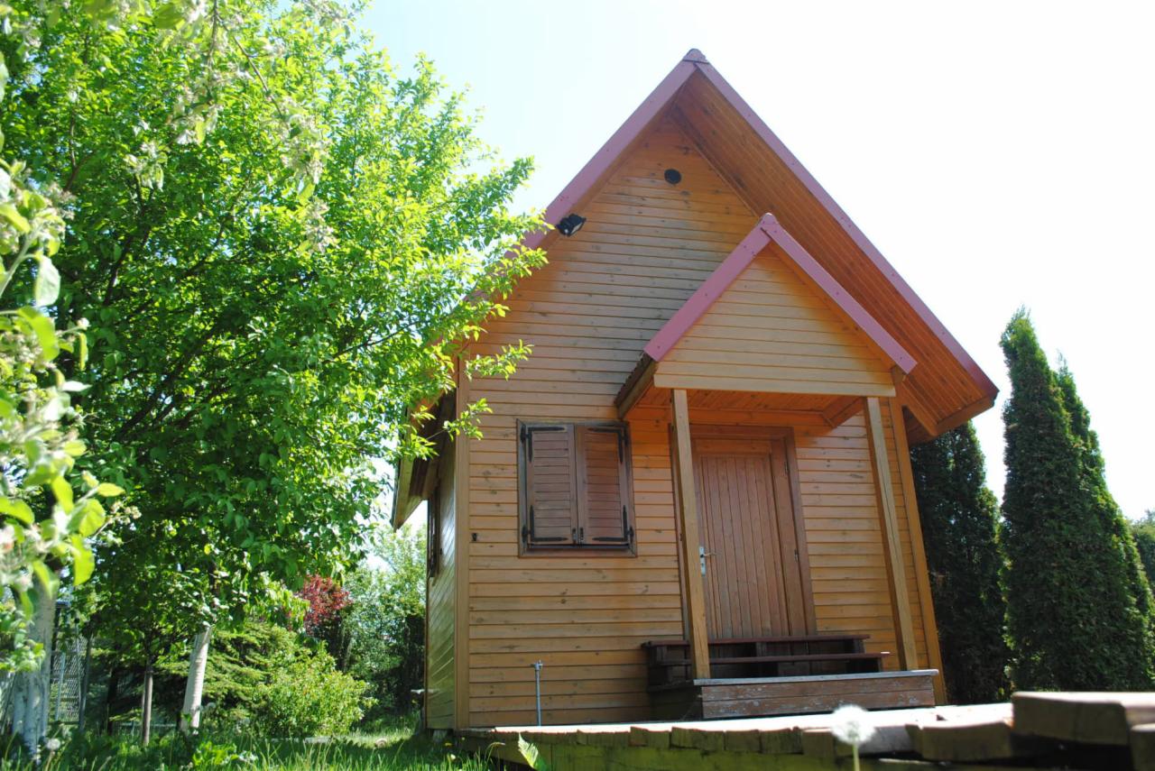Niski, drewniany dom szkieletowy z czerwonym dachem, otoczony zielenią drzew i krzewów. Widoczny fragment tarasu i zamknięte okiennice.