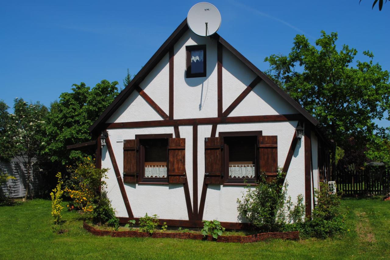 Biały dom szkieletowy z brązowymi elementami konstrukcyjnymi, drewnianymi okiennicami i anteną satelitarną na dachu, otoczony zielenią.