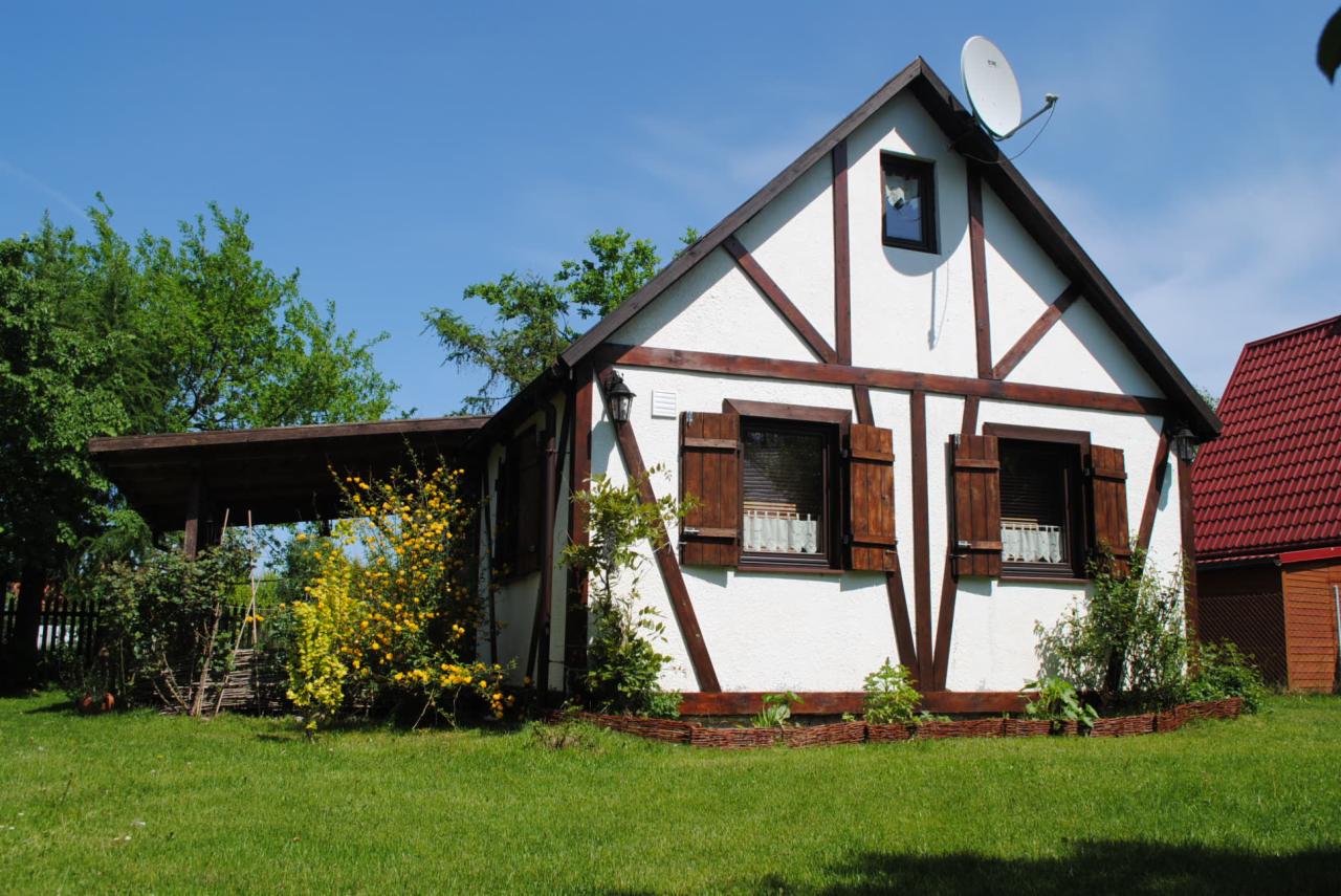 Biały dom szkieletowy z brązowymi belkami i okiennicami, widoczny z boku, otoczony zielenią i niskim płotkiem z wikliny, z zadaszeniem po lewej stronie.