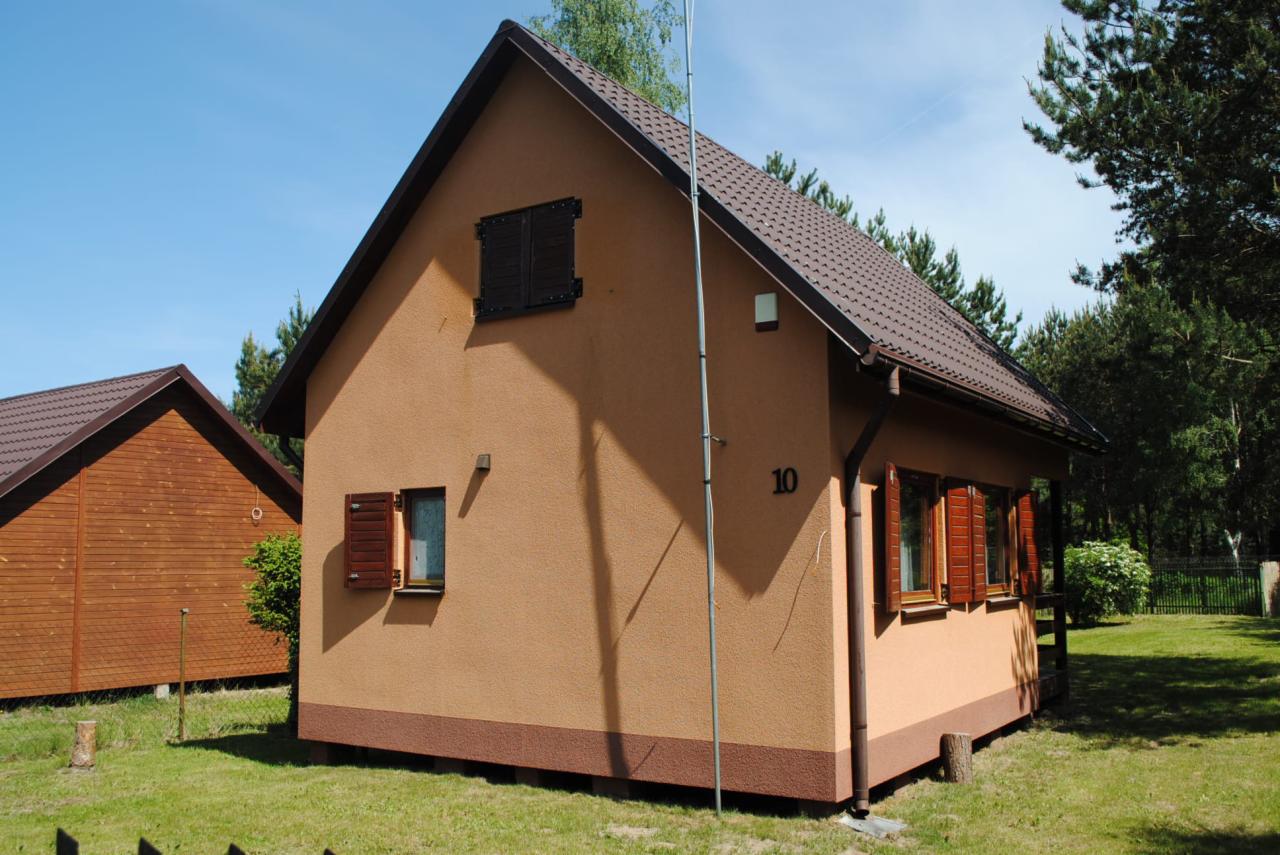 Niewielki, parterowy dom z beżową elewacją, brązowym dachem i okiennicami, numerem '10' na ścianie. Obok widoczny drugi, drewniany budynek. W tle drzewa i błękitne niebo.