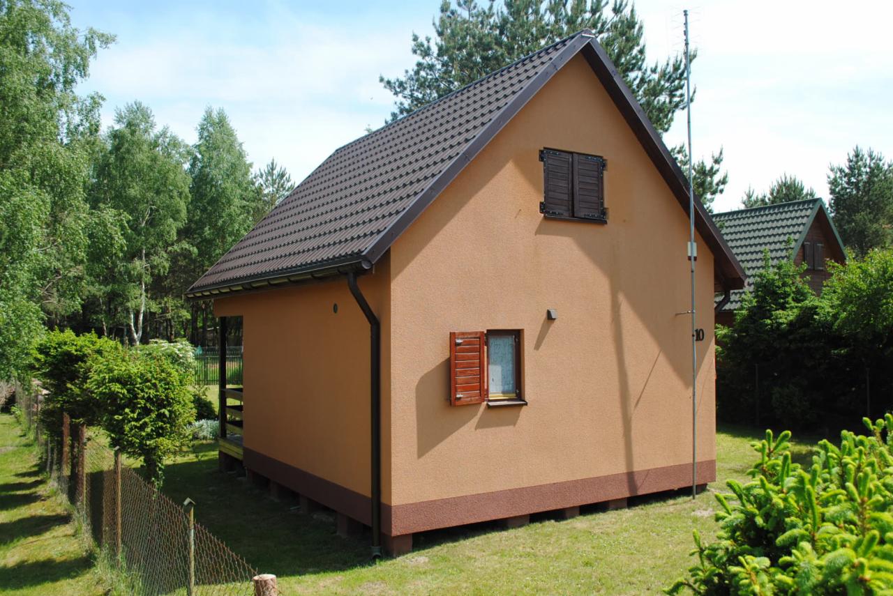 Niewielki, parterowy dom szkieletowy z brązowym dachem i drewnianymi okiennicami, otoczony zielenią. Widoczna antena na dachu i numer domu '10'.