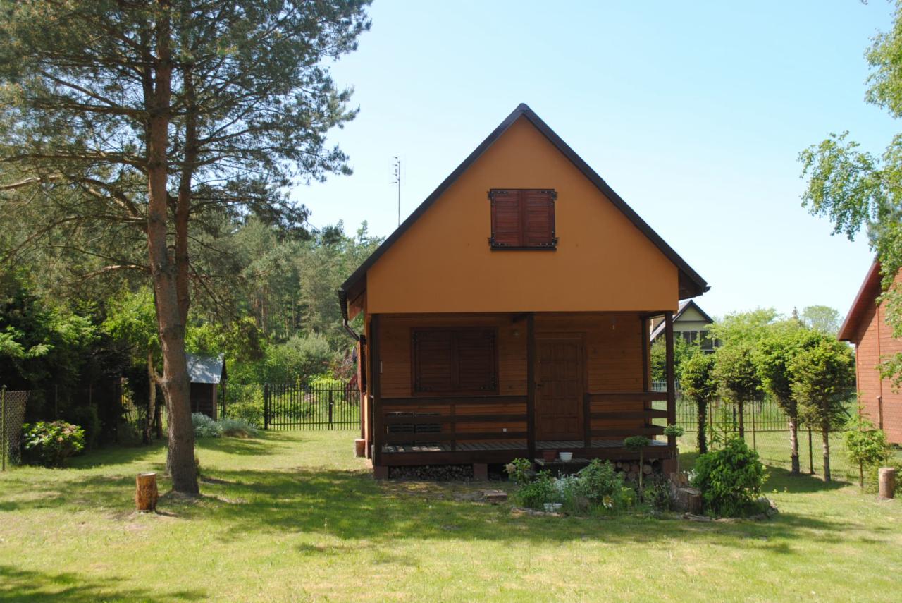 Uroczy, parterowy dom z drewnianą werandą i okiennicami w odcieniach brązu, otoczony zielonym ogrodem z drzewami i krzewami. Na dachu widoczna antena.