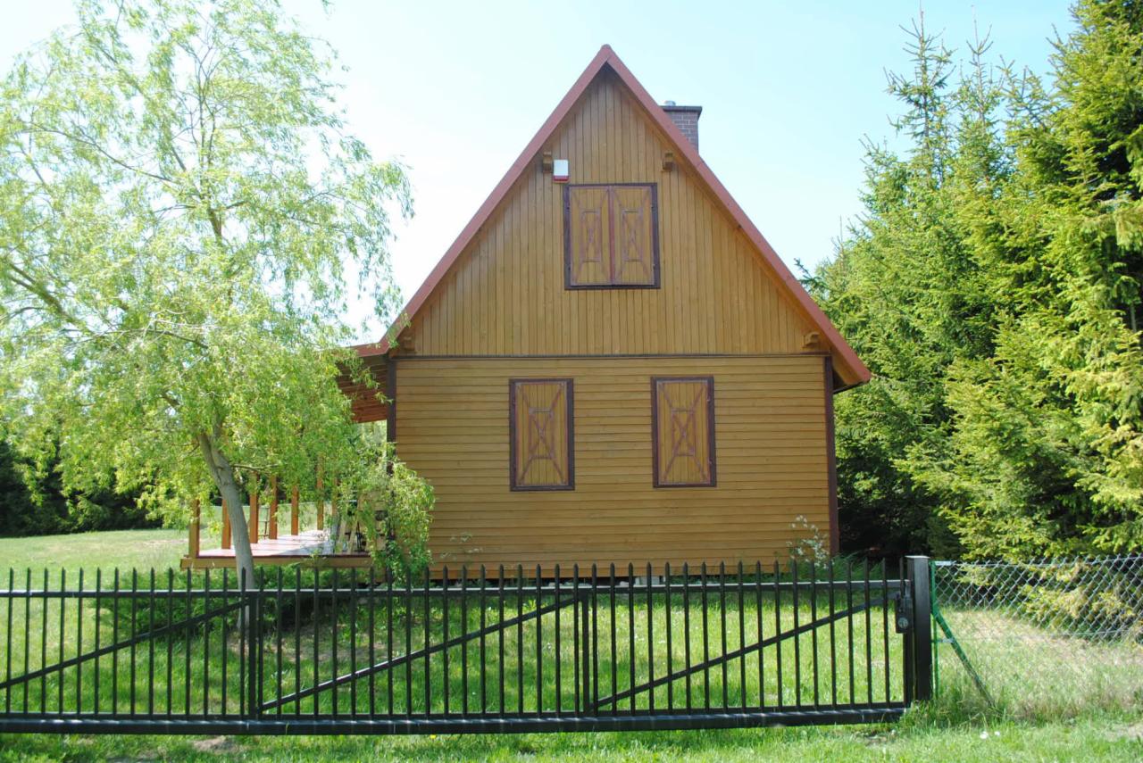 Żółty dom szkieletowy z brązowymi okiennicami za czarnym ogrodzeniem, widok zza ogrodzenia, drzewa w tle. Dom z poddaszem i kominem.