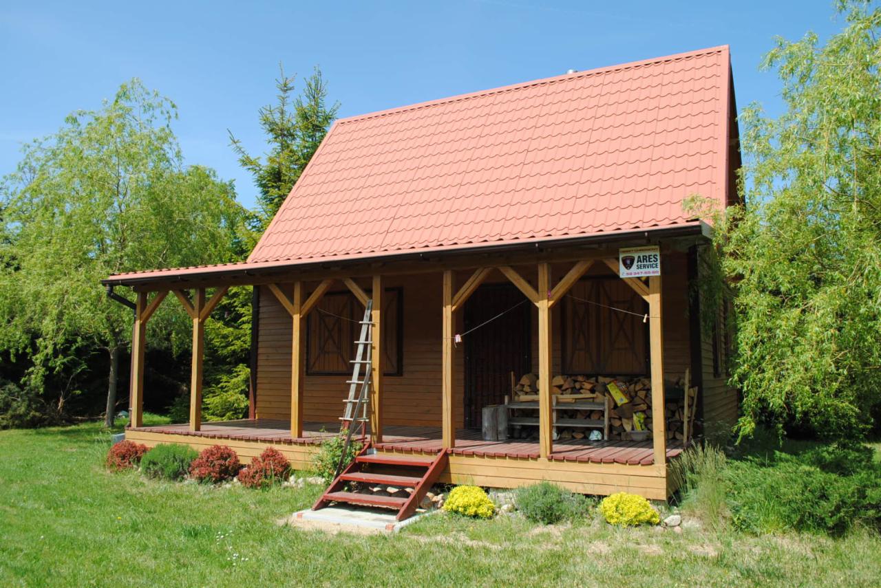 Drewniany domek letniskowy z zadaszonym tarasem, schodami wejściowymi i widocznym drewnem na opał. Czerwony dach z dachówki. Na elewacji widoczny szyld firmy.