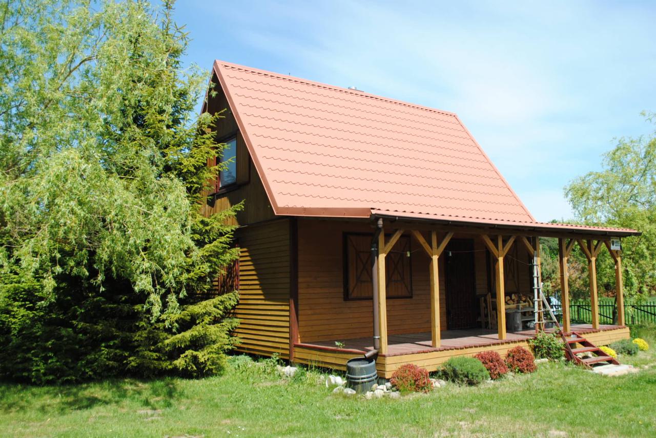 Drewniany dom szkieletowy z gankiem i pomarańczową blachodachówką, otoczony zielenią. Widoczna rynna spustowa i drewniane okiennice. Ujęcie w słoneczny dzień.