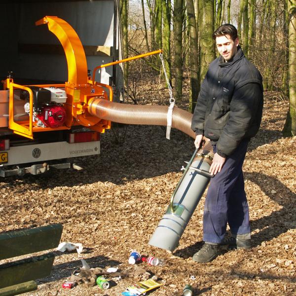 Mężczyzna obsługuje pomarańczową maszynę do rozdrabniania gałęzi zamontowaną na samochodzie VW, sprzątając teren z liści i śmieci w lesie.