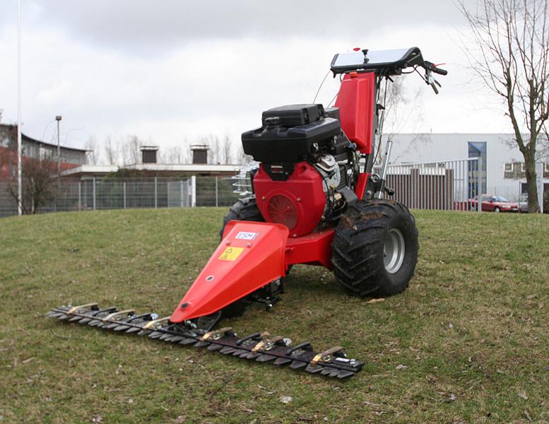 Czerwona kosiarka listwowa ESM na trawiastym wzgórzu, z budynkiem w tle. Widoczne detale: silnik Honda, opony z bieżnikiem, listwa tnąca.