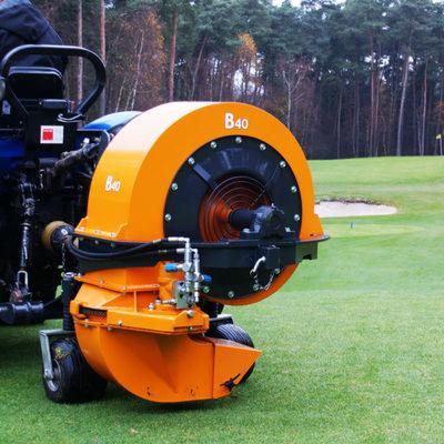 Pomarańczowa maszyna B40 do pielęgnacji trawy na tle zielonego pola golfowego i drzew. Widoczny fragment operatora i maszyny.