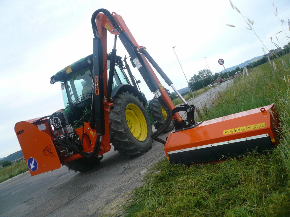 Pomarańczowy ciągnik z kosiarką listwową na poboczu drogi, widok z boku, z żółtymi kołami i zieloną kabiną, w tle krajobraz z drogą i znakami drogowymi.