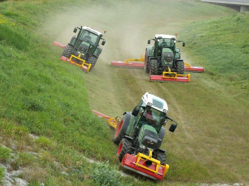 Trzy traktory koszą trawę na zboczu wzgórza. Zielone maszyny z czerwonymi i żółtymi elementami pracują w trudnym terenie, tworząc równo przyciętą powierzchnię.